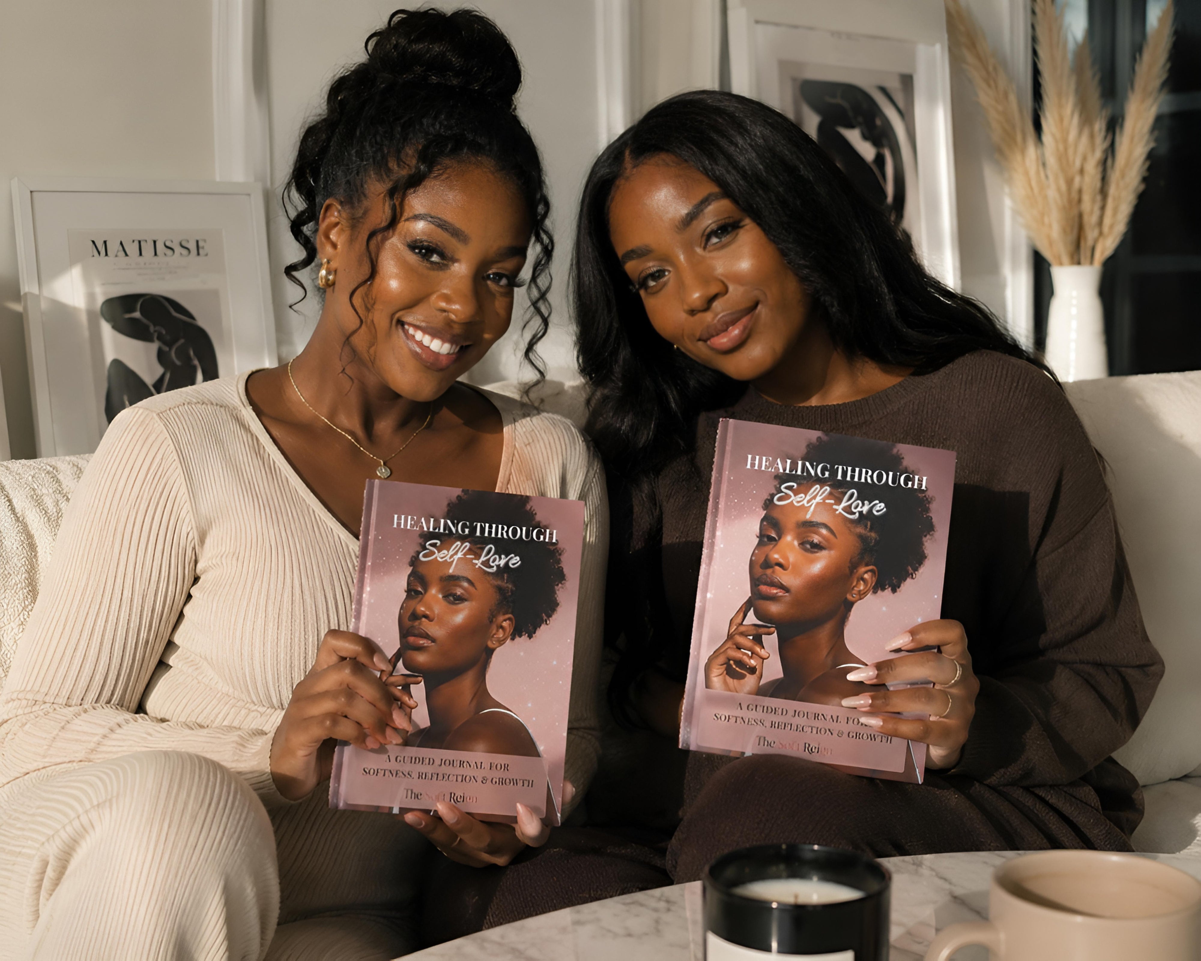 Two women sitting on a couch indoors, each holding a copy of the book 'Healing Through Self Love' by Claudia Gray. The books have pink covers with a portrait of a Black woman and white text.